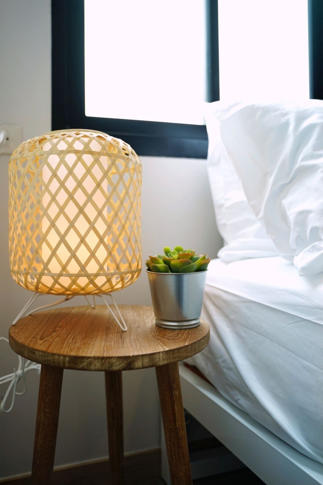 A bed with white linens, a wooden stool with a metal lamp, and a small potted succulent plant.