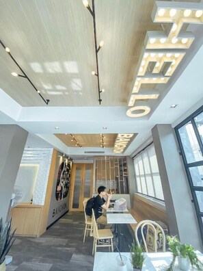 A modern interior with a ceiling featuring illuminated letters spelling "HELLO" and "BANGKOK". There is a person seated at a table with a laptop, and the room has large windows and a wooden cabinet in the background. - YakThai Poshtel (Bangkok)