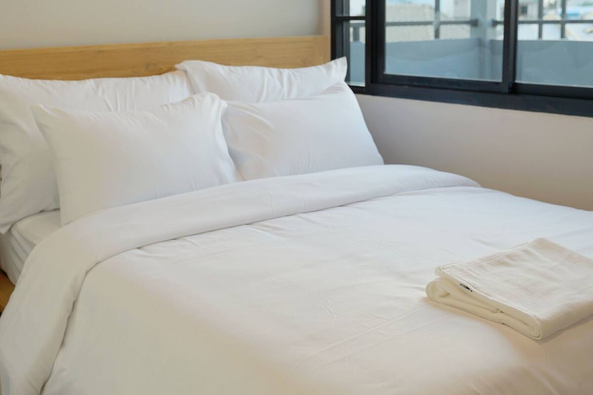 A neatly made bed with white linens in a room with a large window.