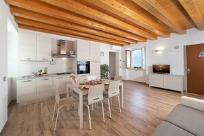 Et moderne køkken med hvide skabe, et spiseområde med et træbord og stole, samt en opholdsstue med et fjernsyn.
