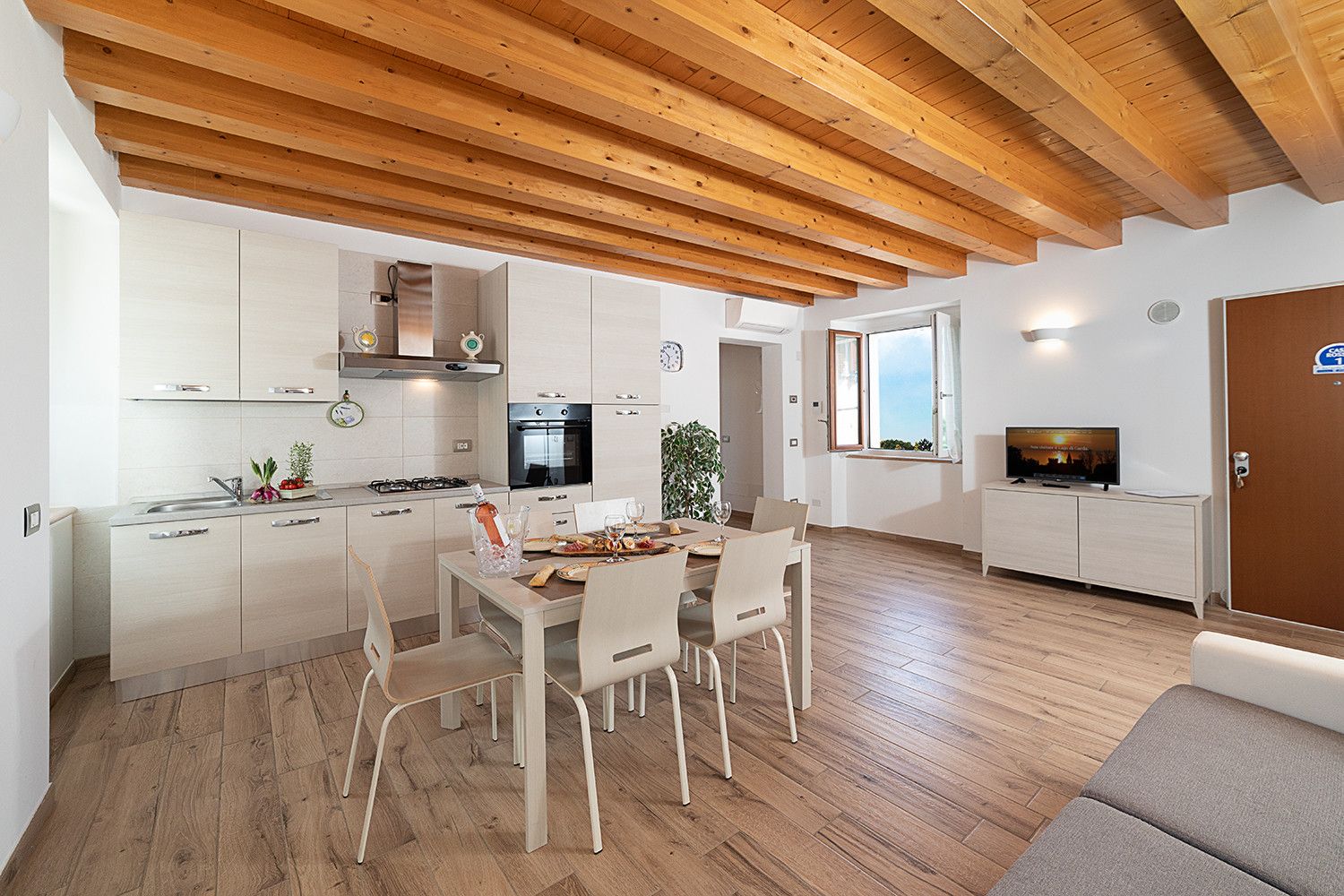 A modern kitchen with white cabinets, a dining area with a wooden table and chairs, and a living area with a television and a sofa.