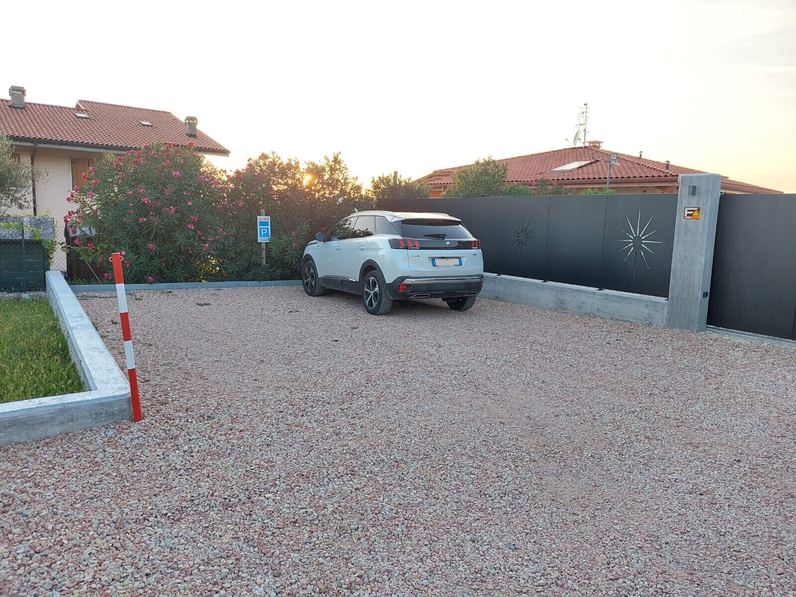 Een witte SUV staat op een grindpad met een hekpoort, een parkeerbord en een decoratieve muur met een sterrenmotief.