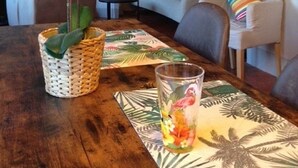Zona pranzo con tavolo in legno, cesto intrecciato con una pianta e bicchiere con motivo tropicale.