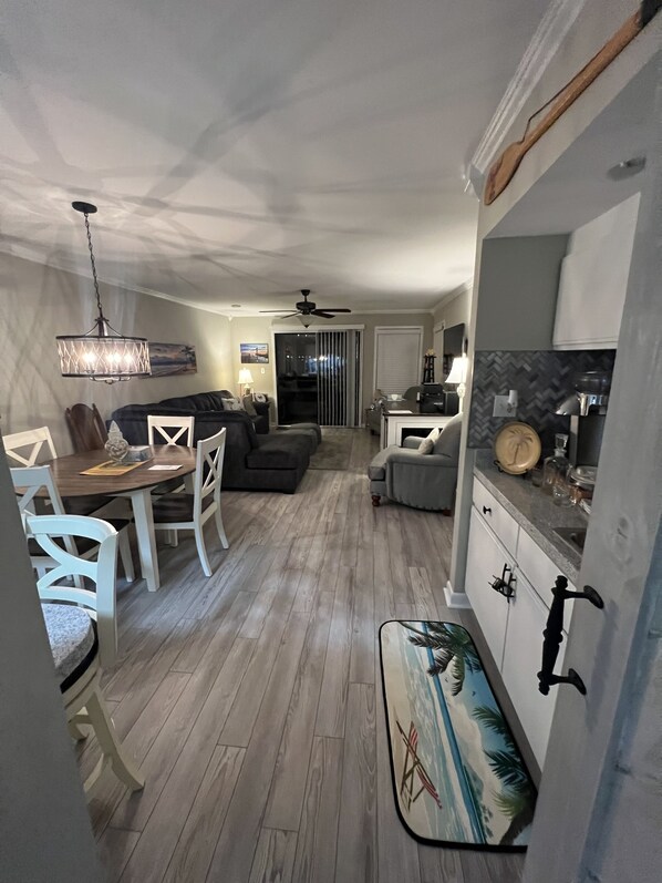 Una cocina moderna con armarios blancos, un comedor con mesa y sillas de madera, y una sala de estar con sofá gris y ventilador de techo.