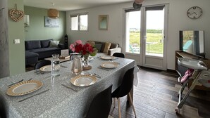 Comedor con mesa lista para cuatro, un sofá al fondo y vista a través de la puerta de cristal.