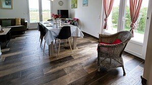 Amplia sala de estar con una mesa de comedor, sillas, un sofá y una silla de mimbre. Hay un televisor, un reloj y una ventana con cortinas.