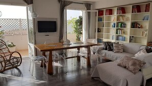 A living room with a wooden dining table, clear chairs, a white sofa, and a bookshelf filled with books.