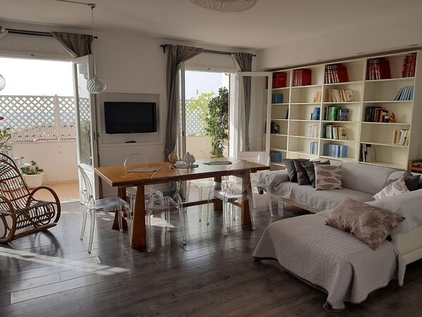 A living room with a wooden dining table, clear chairs, a white sofa, and a bookshelf filled with books. - Miguel Apartment located in the center of Tropea, a stone's throw from the sea (Tropea)