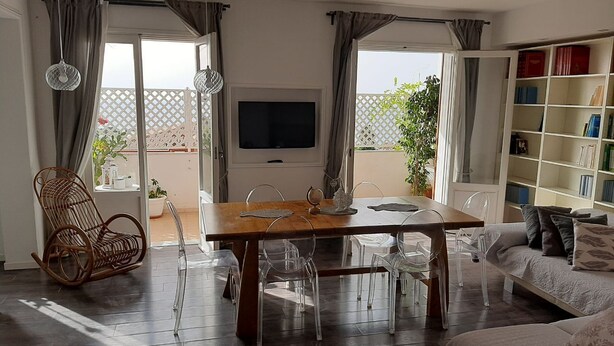 A modern living room with a wooden dining table, clear chairs, a wicker chair, a flat-screen TV, a bookshelf, and a sofa.