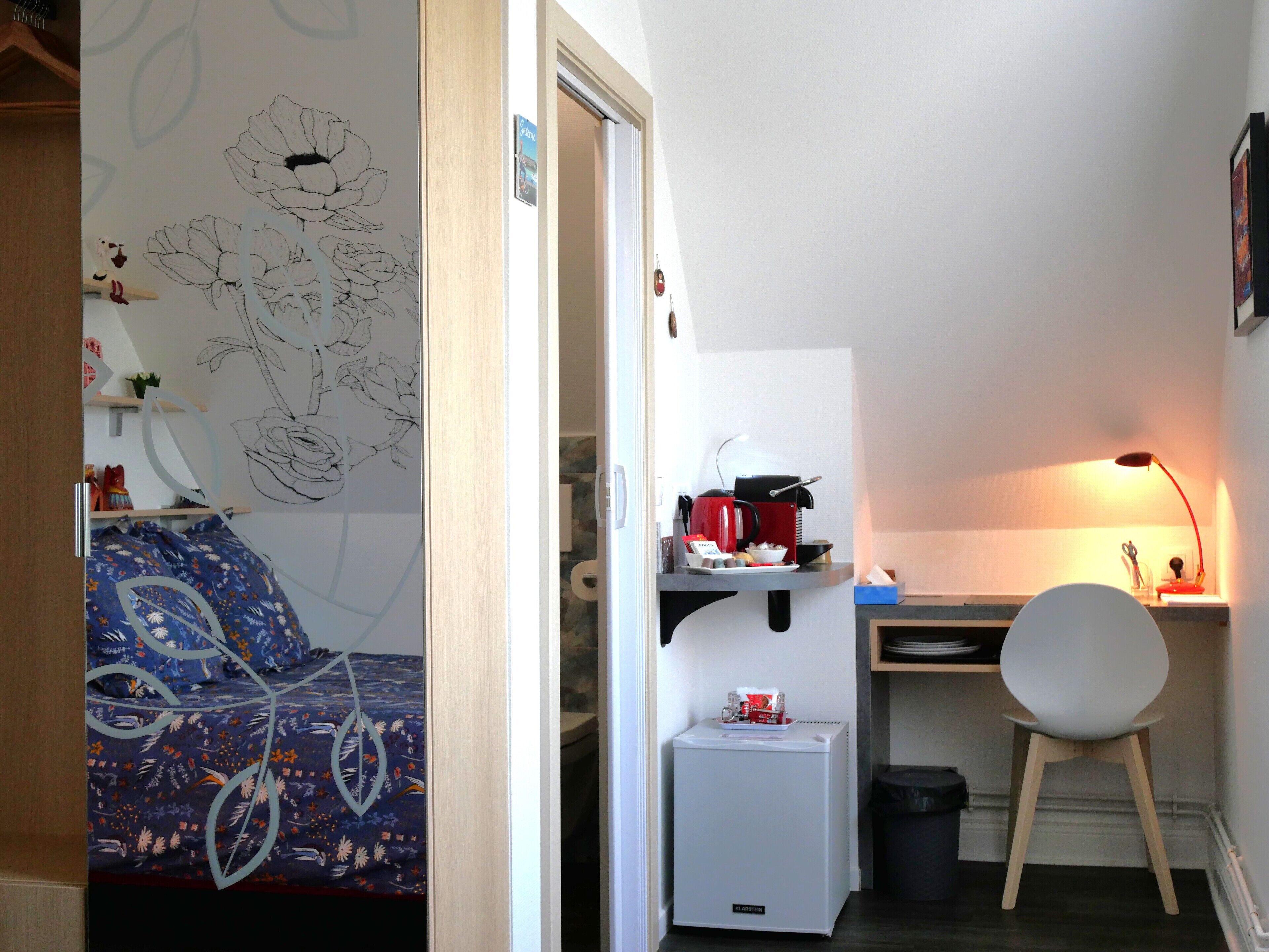 A compact room with a bed, a desk, a chair, a small refrigerator, and a trash bin.