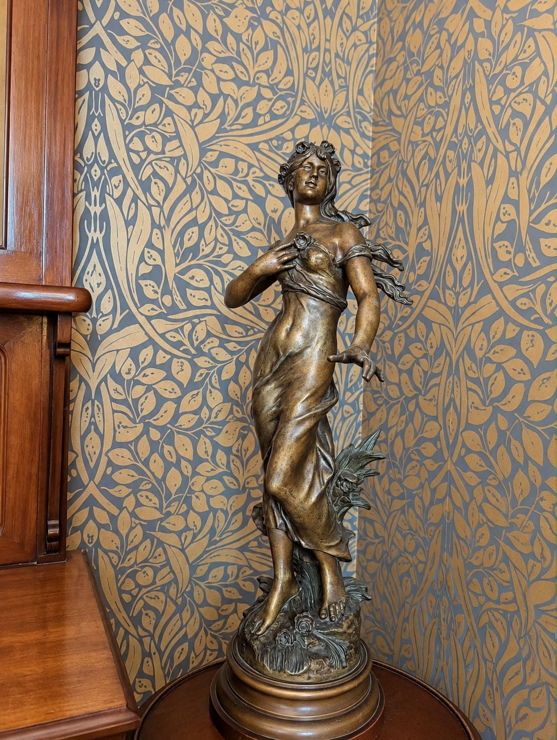 Una estatua de bronce de una mujer sosteniendo una flor, sobre un pedestal de madera, con un fondo de papel tapiz estampado.