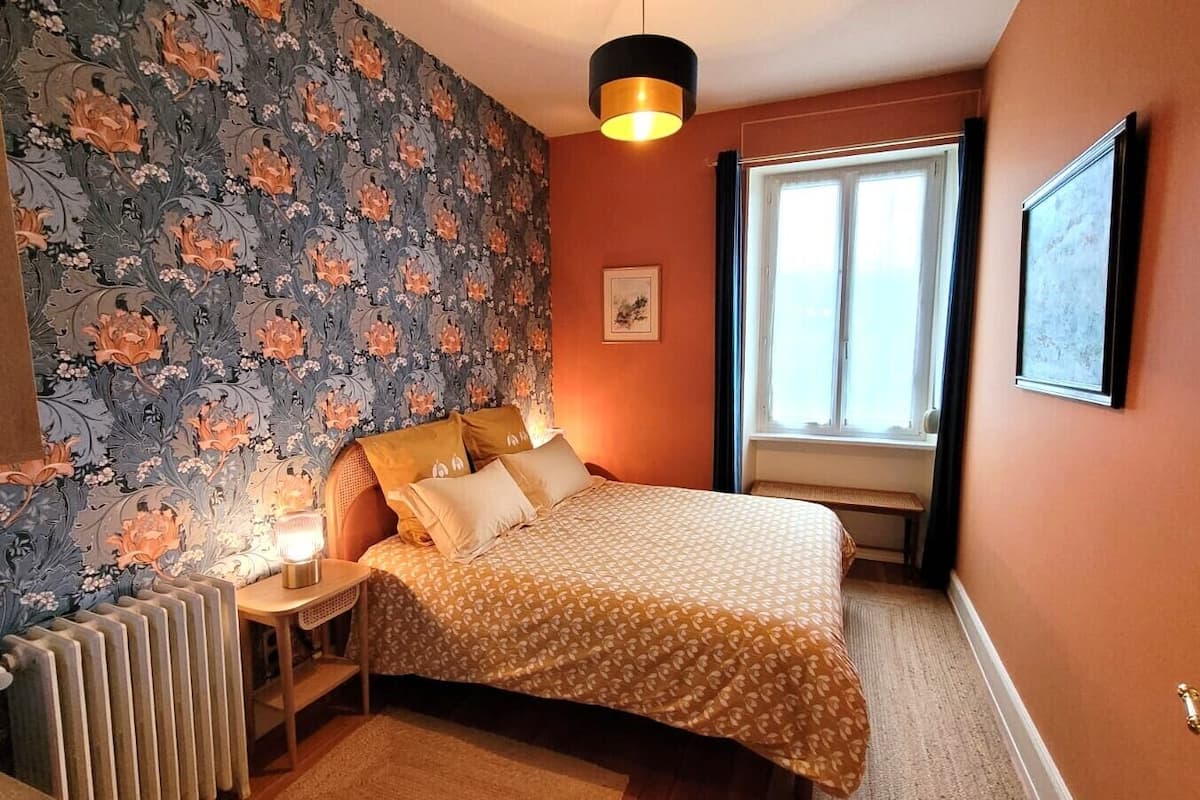 Ein Schlafzimmer mit Bett, Nachttisch, Heizkörper, einem Fenster mit Vorhängen und einem Wandbild.