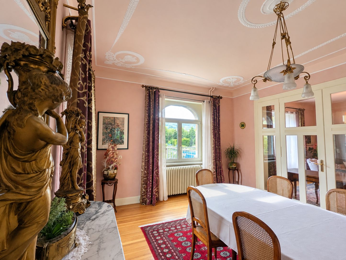 Een eetkamer met een marmeren tafel, rieten stoelen, een rood tapijt met patroon en een beeld van een vrouw.