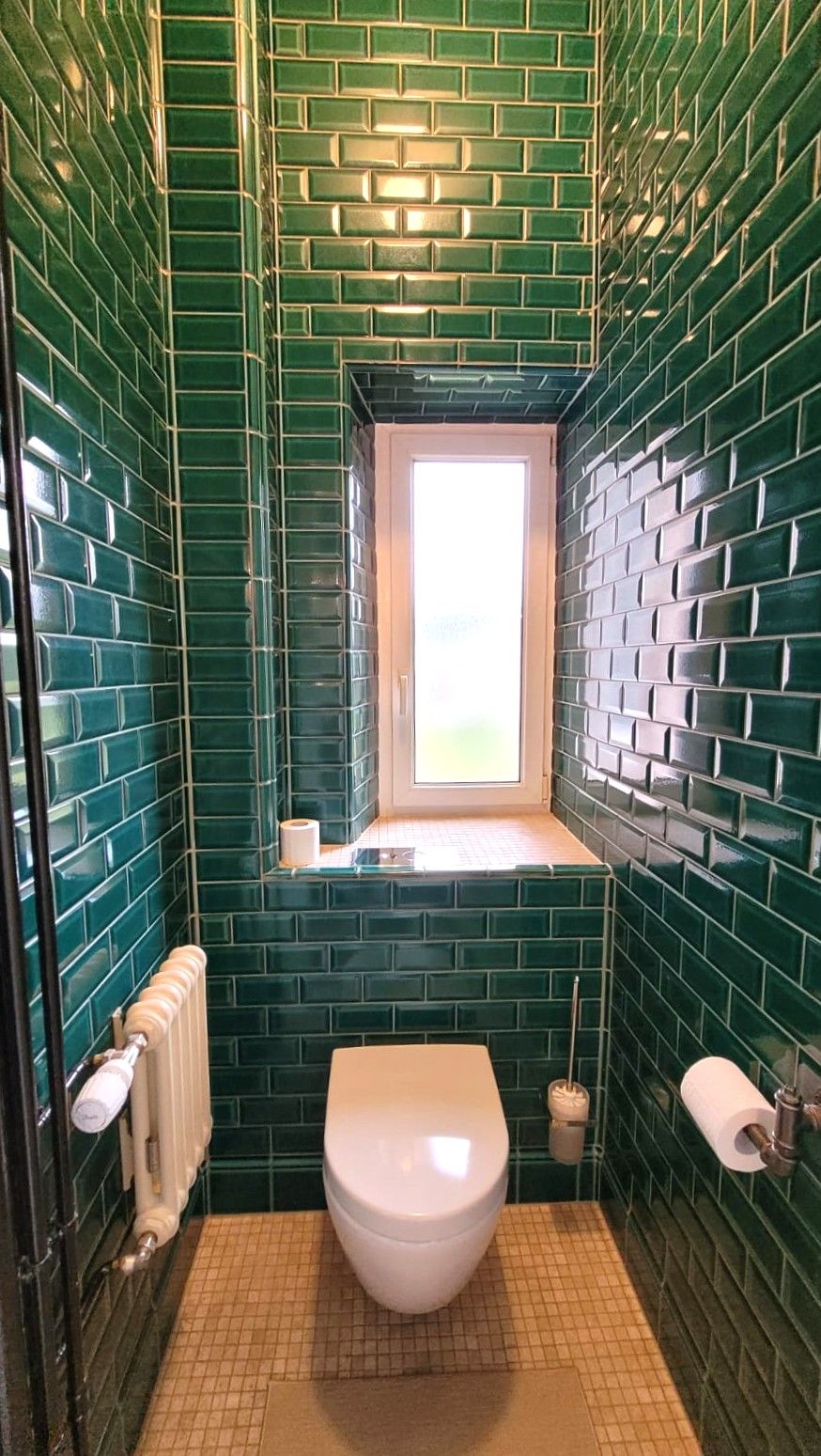 Un baño con paredes de azulejos verdes, un inodoro blanco, una ventana y un perchero para toallas.