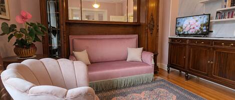 A living room with a pink sofa, a matching armchair, a wooden cabinet, a television, and a patterned rug.
