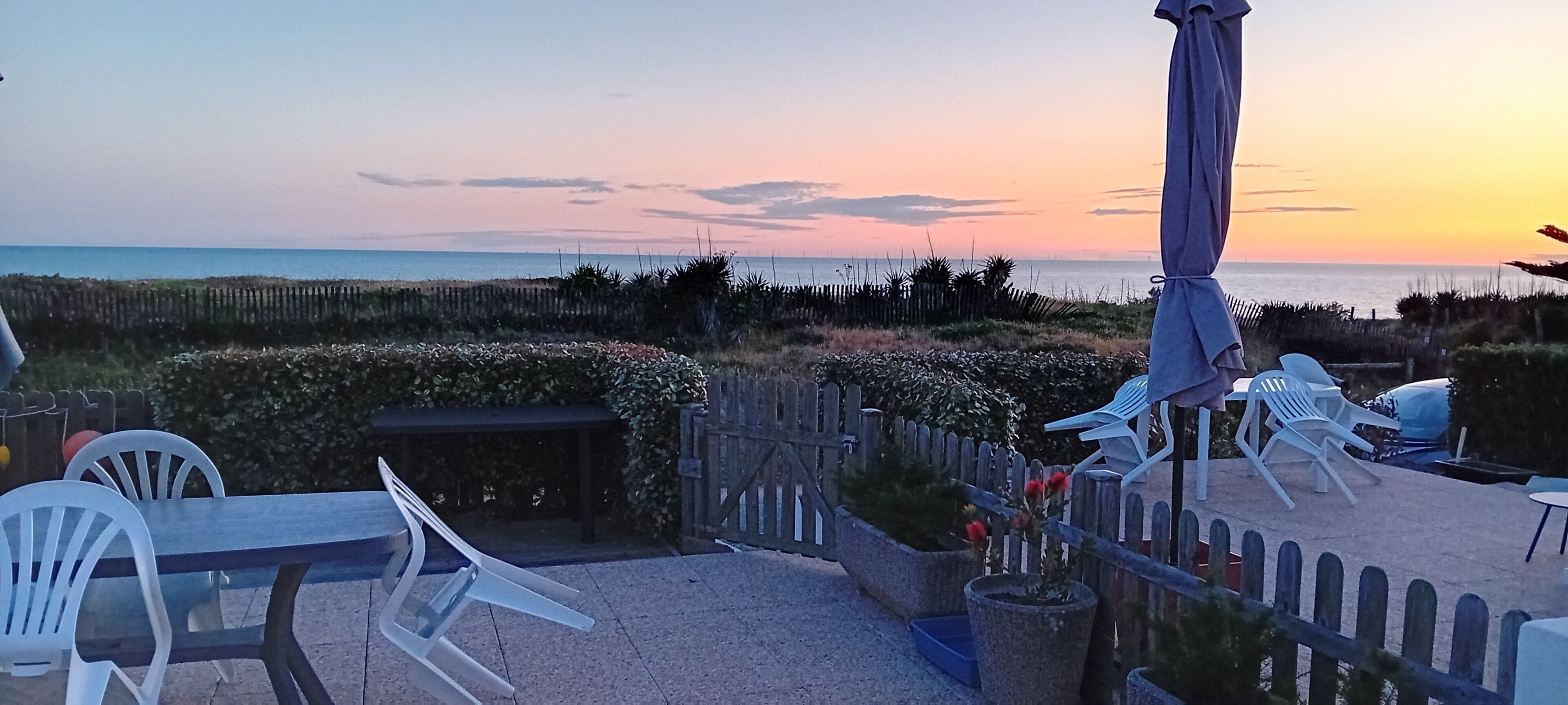 Une terrasse avec des meubles d’extérieur, une clôture en bois et une vue sur l’océan au coucher du soleil.