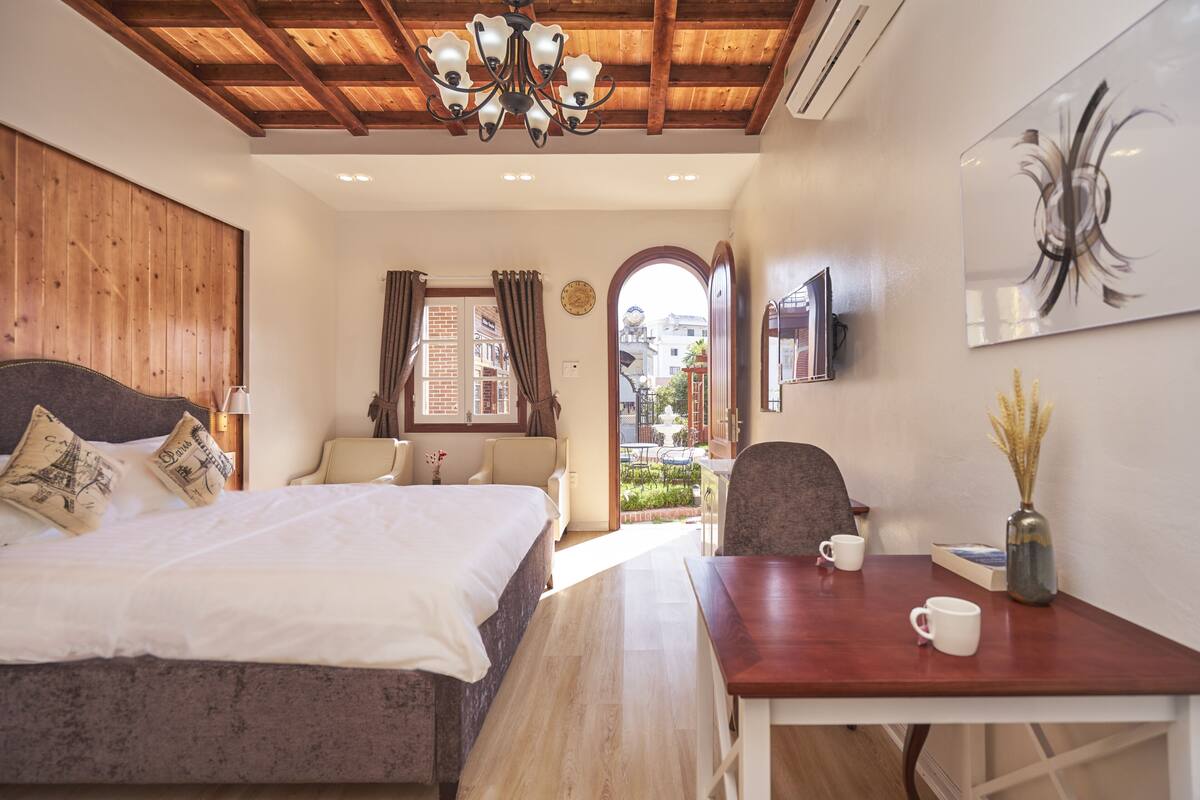 A bedroom with a bed, a desk, a chair, a vase with dried plants, and a view of the outside.