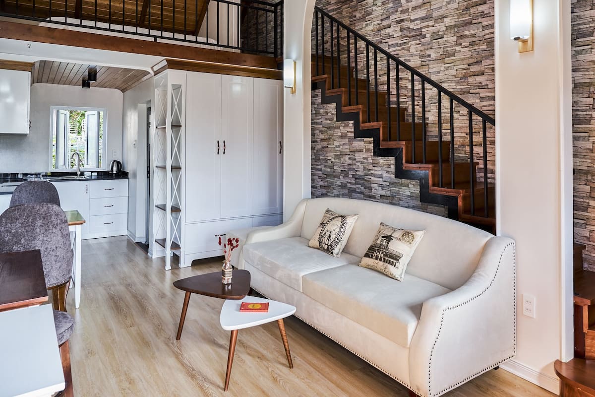 A spacious living area with a high ceiling, a staircase with a stone wall, and a white sofa with decorative pillows.