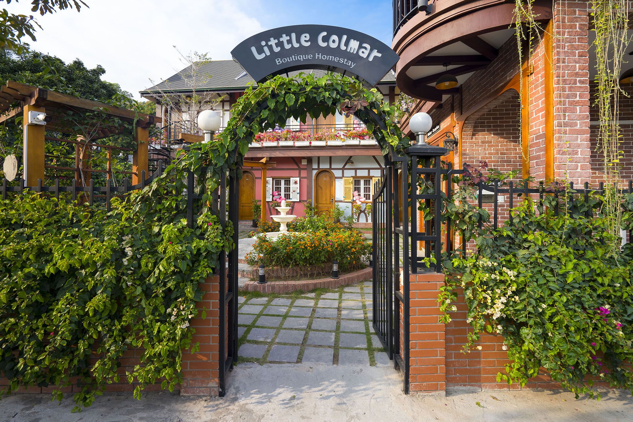 A boutique homestay entrance with a sign reading "Little Colmar Boutique Homestay," a brick building, and a garden area with a fountain.