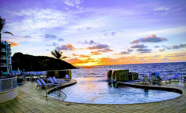A poolside area with lounge chairs and a view of the sunset over the ocean.