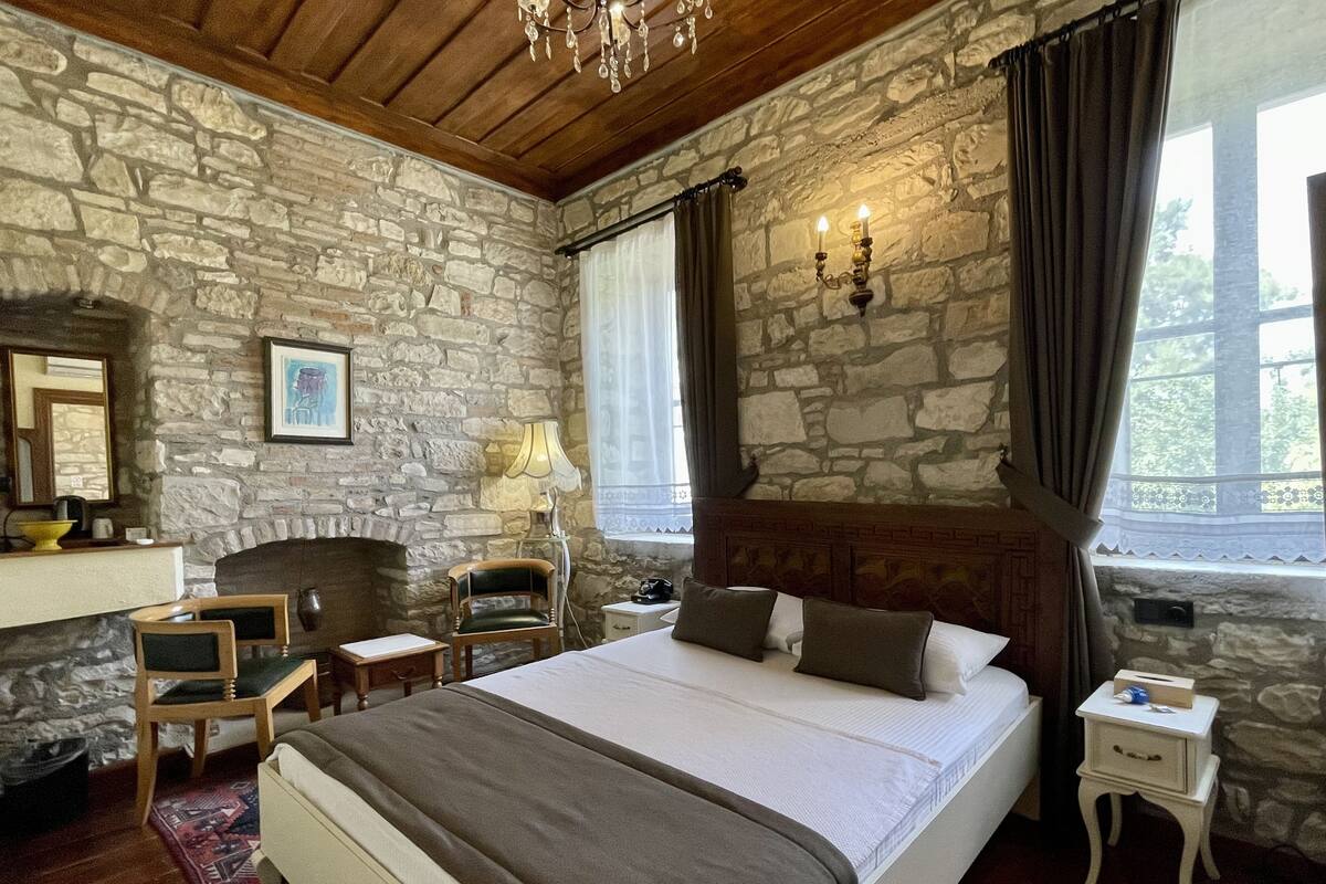 A bedroom with a stone wall, a wooden ceiling, a bed with pillows, a nightstand, a lamp, and a window with curtains.