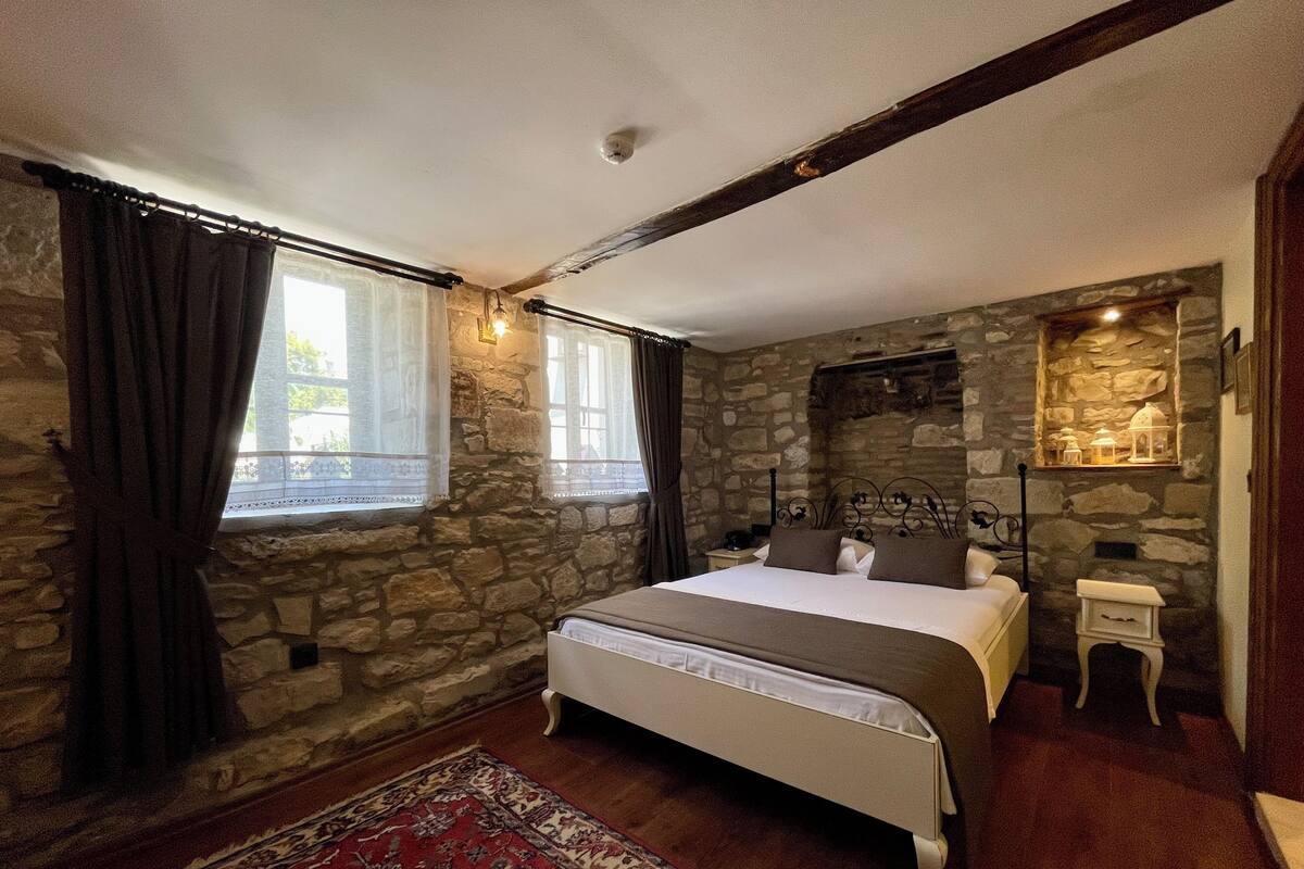 A bedroom with a stone wall, a bed with white linens, a nightstand, a red patterned rug, and a stone shelf with decorative items.