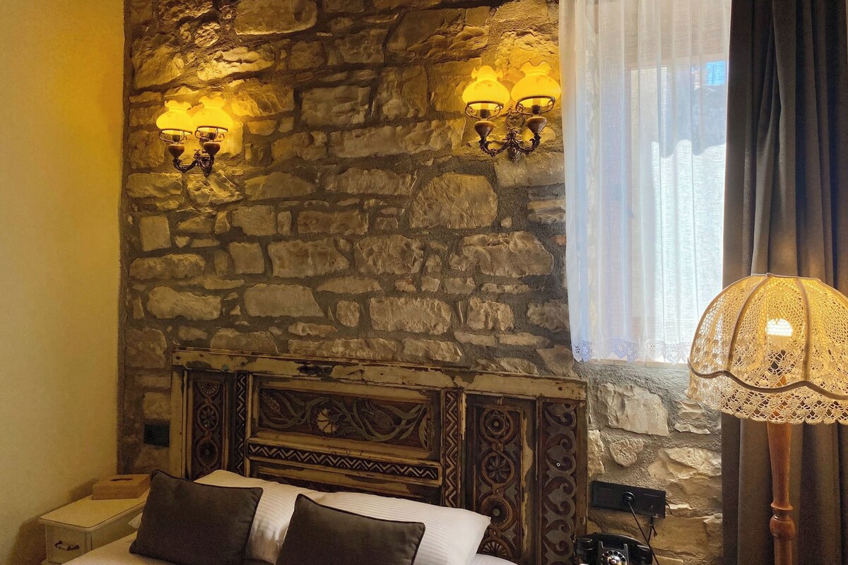 A bedroom with a stone wall, a bed with a headboard, two wall sconces, a window with sheer curtains, and a decorative lamp.