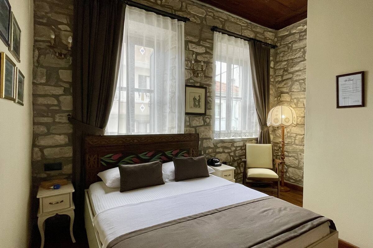 A bedroom with a stone wall, a wooden ceiling, a bed with white bedding and two pillows, a nightstand with a lamp, and a chair.