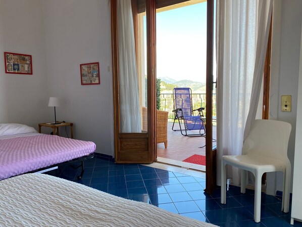 A hotel room with a bed, a chair, a small table with a lamp, and a balcony with a chair.