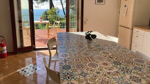 Eine Küche mit Fliesenboden, einem Tisch mit Stühlen, einem Kühlschrank und Meerblick durch eine Glastür.