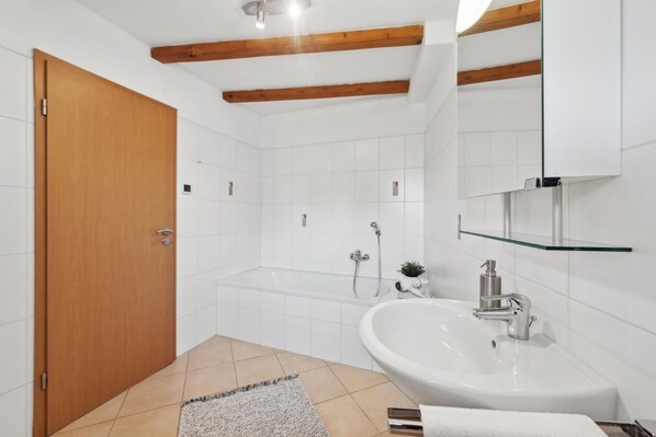 A modern bathroom with a wooden door, white tiles, a glass shelf, a sink, and a shower.