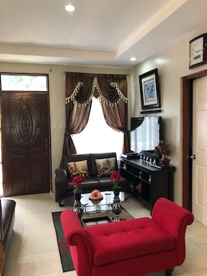 A living room with a brown door, a brown sofa with decorative pillows, a red sofa, a coffee table with flowers, and a black entertainment unit.
