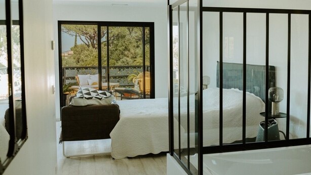 A bedroom with a large window, a bed, a nightstand, and a bathroom with a bathtub.