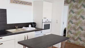 A modern kitchen with white cabinets, a dark countertop, stainless steel appliances, and a table with metal legs. - House with enclosed garden, on the sea side (Brem-sur-Mer)