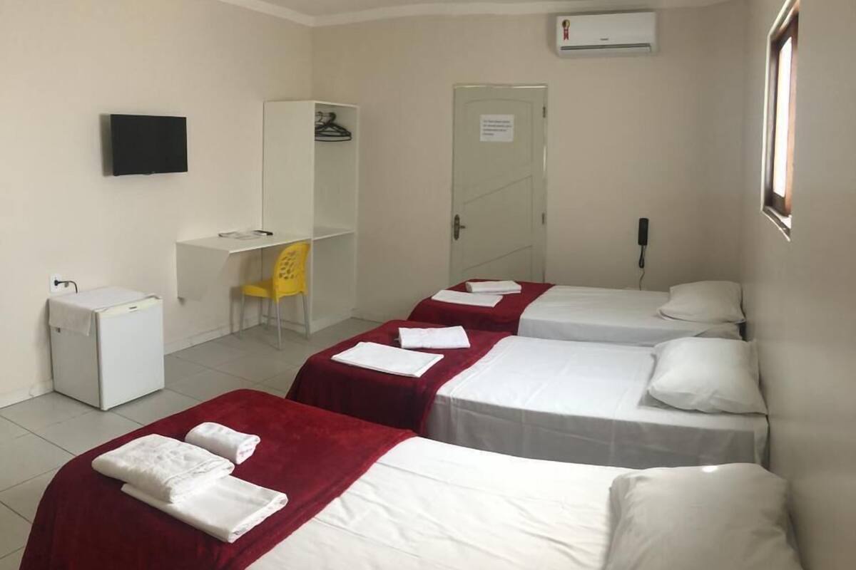 A hotel room with three single beds, a desk, a chair, a wardrobe, an air conditioning unit, and a TV.