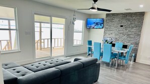 Un salon moderne avec un canapé d’angle gris, un coin repas avec des chaises bleues, un téléviseur fixé sur un mur en pierre et de grandes fenêtres donnant sur une terrasse en bois.