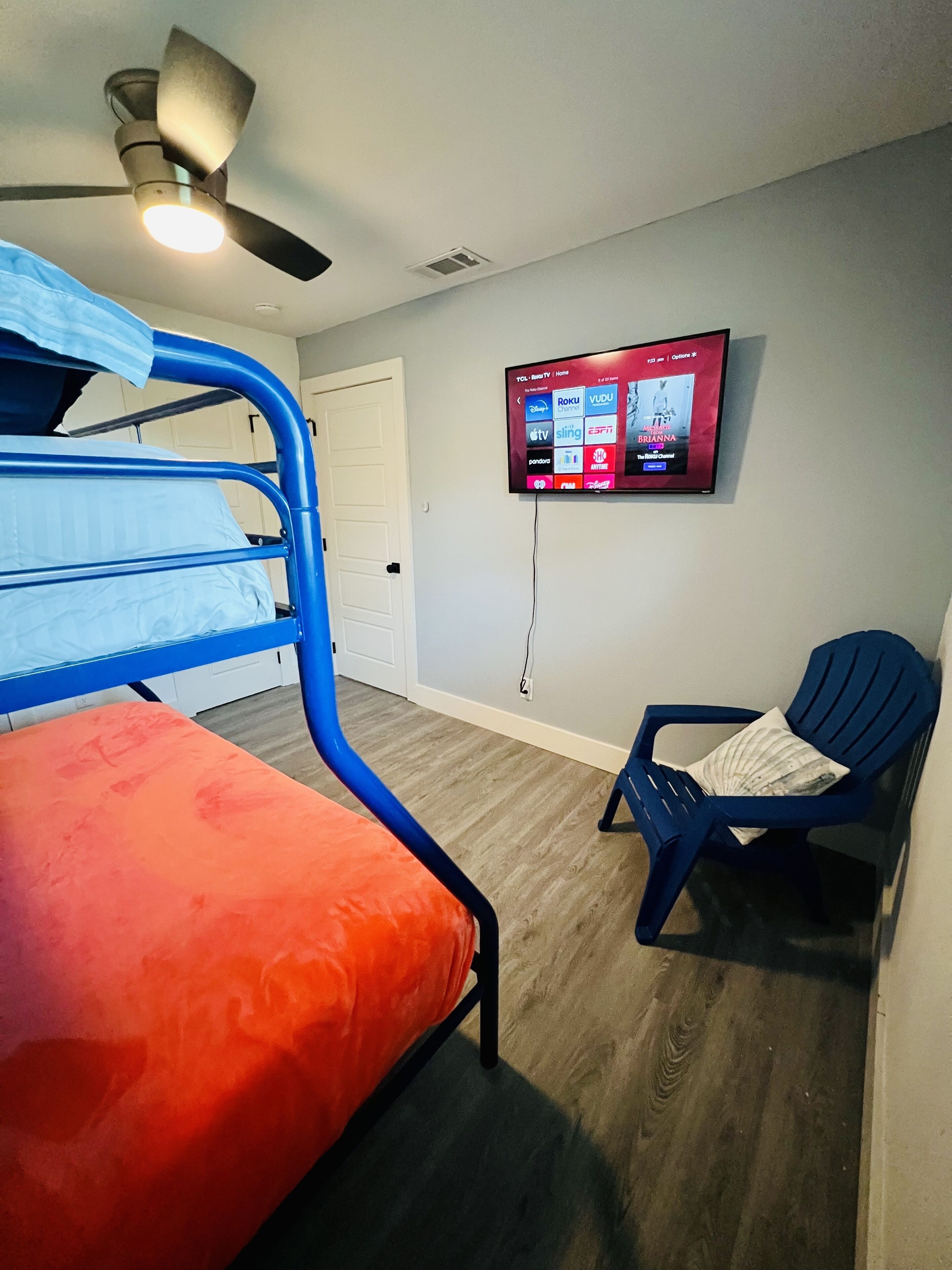 Une chambre avec des lits superposés, un ventilateur de plafond, un téléviseur fixé au mur et une chaise bleue.
