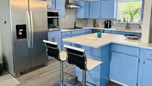 Une cuisine moderne avec des armoires bleues, un réfrigérateur en acier inoxydable, un four encastré et une machine à café. On y trouve deux tabourets de bar et une fenêtre agrémentée de plantes.