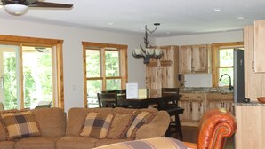 A spacious living room with a brown leather sofa, a plaid armchair, and a dining area with wooden cabinets and a chandelier. - 3BR Cottage Nestled in a Door County Serene Setting (Sturgeon Bay)