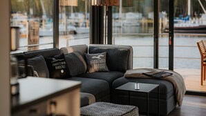 Ett modernt vardagsrum med en soffa, ett litet bord och utsikt över en småbåtshamn genom stora fönster.