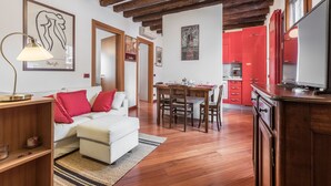 A modern living room with a white sofa, red cushions, a wooden coffee table, and a lamp. There is a dining area with a wooden table and chairs, and a kitchen with red cabinets and a large window.