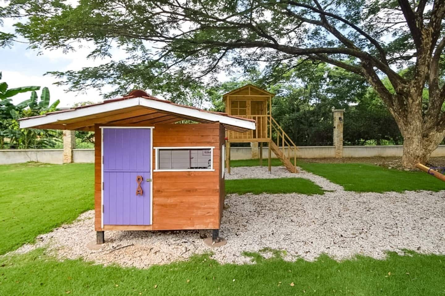 A small wooden playhouse with a purple door and a slide, surrounded by a grassy area with gravel paths.