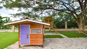 Una casita de juegos de madera con una puerta morada y un tobogán, rodeada de césped y caminos de grava.