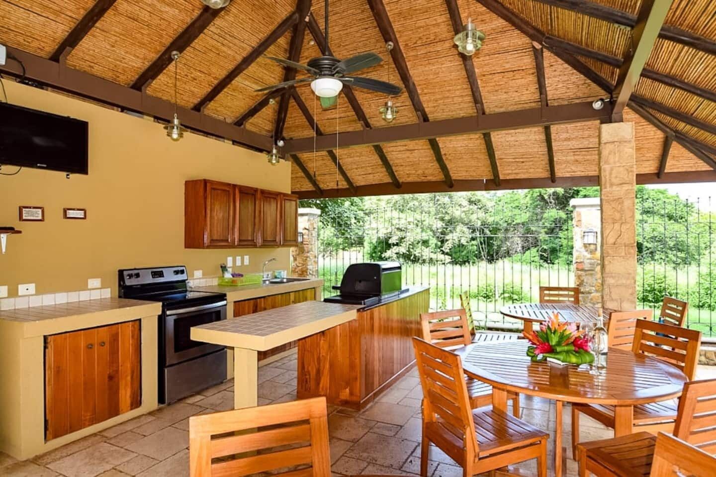 A covered outdoor kitchen with a dining area, including a table and chairs, a ceiling fan, and a view of the surrounding greenery.