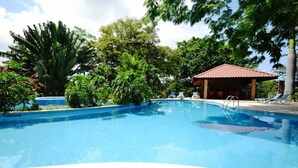 Una piscina con borde de baldosas, rodeada de abundante vegetación y un pequeño pabellón con techo rojo.