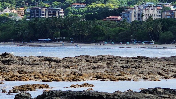 Zona costera con costa rocosa, edificios en una ladera y una playa con gente.