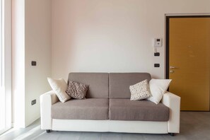 A neutral-colored sofa with two pillows, a door, and a wall-mounted control panel.