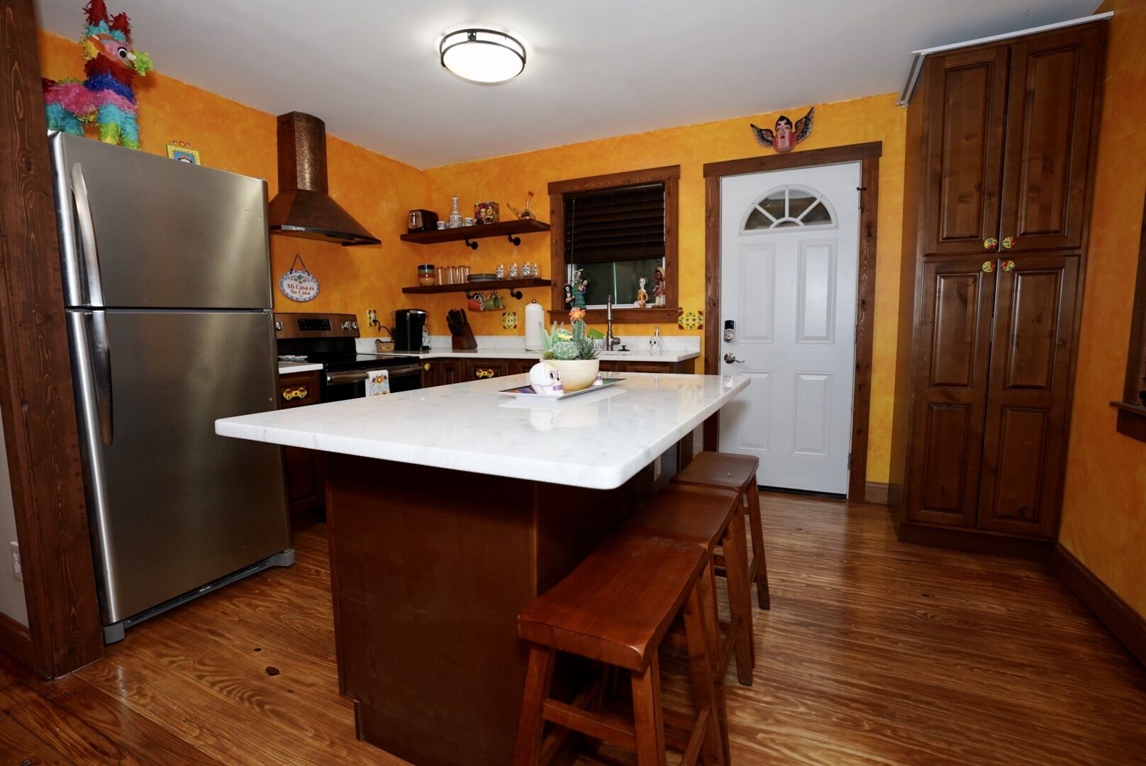A kitchen with a stainless steel refrigerator, wooden cabinets, a marble countertop, and a wooden bench.