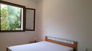 Quarto com cama de madeira, mesa de cabeceira com luminária, pequena mesa e janela com vista para árvores.