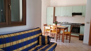 Uma sala de estar com sofá e uma mesa de jantar com cadeiras. Há uma área de cozinha com armários e fogão.